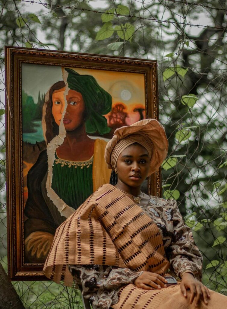 A striking young woman, adorned in traditional Nigerian attire including a matching turban and shawl, sits in front of a framed painting that juxtaposes a torn image of the Mona Lisa with a portrait of a Black woman wearing a green headwrap. The painting, which appears to be hanging on a chain-link fence intertwined with leafy vines, serves as a backdrop for the modern African art portrait. The woman in the foreground looks directly at the viewer with a serious expression, her clothing featuring intricate patterns and textures in earth tones. The juxtaposition of the classic Western art with the modern African art elements creates a compelling visual statement.