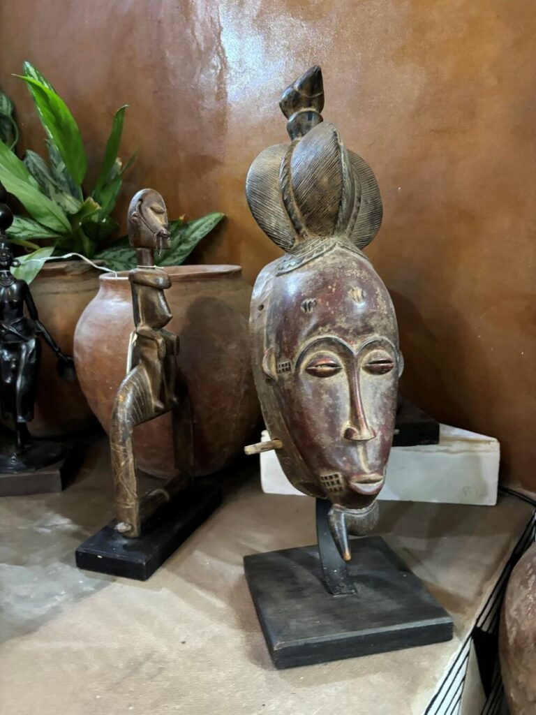 Close-up of a collection of "Traditional African Masks"; a larger face-like mask with a long, narrow nose is on a black base with a long thin support; a smaller statue of a figure kneeling is on a similar black base; also, a small metal statue and a lush green plant in an earthenware pot are in the background.