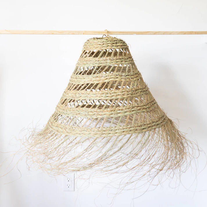 Close-up of a straw hat-shaped lampshade hanging from a wooden beam, against a white wall with an electrical outlet at the bottom. The shade is woven in an open pattern and has a fringe.
