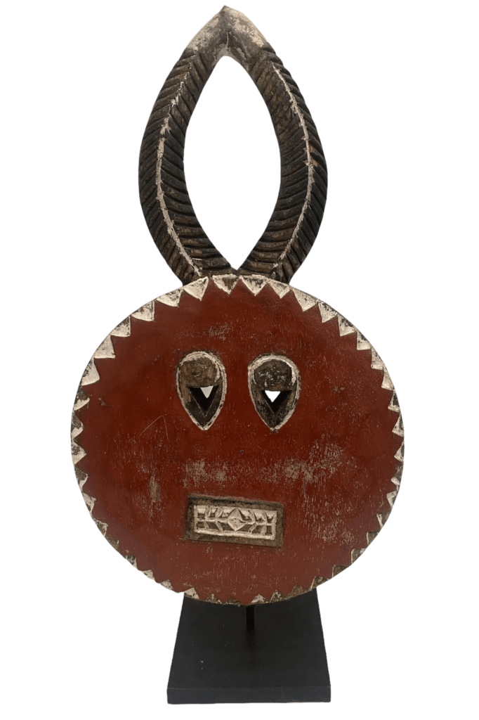 A carved, reddish-brown, oval-shaped, tribal-style mask features two carved eyes and a carved mouth on the surface of the mask and stylized, dark-brown horns extending from the top of the mask, all mounted on a black metal display stand.