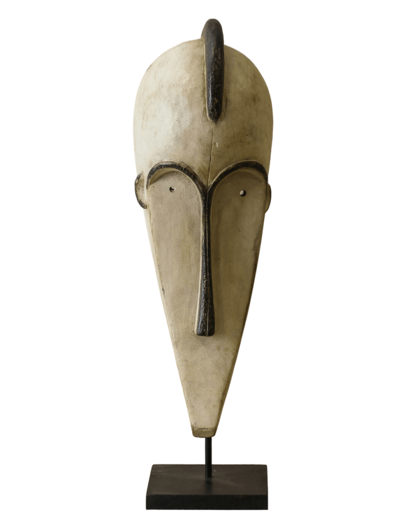 A cream-colored, tribal mask of an elongated human face with a triangular, pointed chin, set atop a narrow, black, square stand.