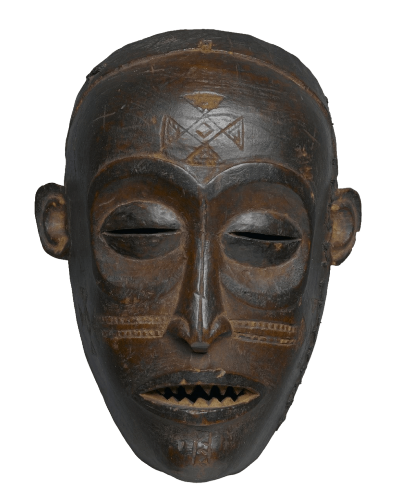 A front view of a brown wooden African mask with decorative carvings against a white background. The mask depicts a human face with elongated features. The eyes are closed, indicated by narrow horizontal slits. Below the nose are carved horizontal lines that resemble a mustache, and the mouth is slightly open to reveal carved sharp teeth. A decorative pattern is carved on the forehead, and small ears are attached to the sides of the mask. The mask appears to be weathered, with visible scratches and variations in tone. It is the central focus of the image, which is well-lit and sharply focused to showcase the mask's details.
