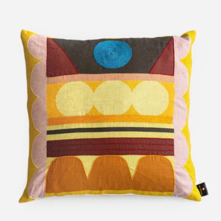 A colorful pillow with a mix of yellow, pink, red, blue and brown hues. It has round and geometric patterns.