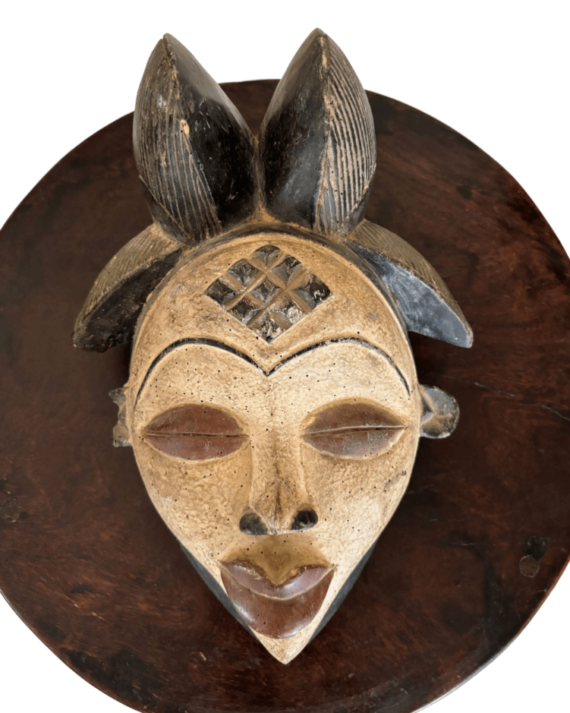 An ornate African ceremonial mask made of dark and light brown wood is mounted on a disk of dark brown wood. The mask has two black, leaf-shaped ornaments at the top, and there is an etched, cross-hatched diamond shape between them. The face and neck of the mask are white with brown details around the eyes and the mouth, which is elongated and triangular. The mask sits in the center of a round, dark brown wooden disk that serves as a backdrop.