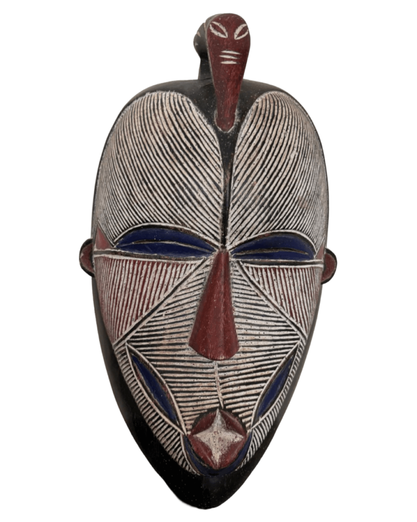 A front view of an elaborately carved and painted wooden tribal mask, likely of African origin. The mask is symmetrical and features a blend of geometric patterns and stylized facial features. The main color is a dark brown or black, which serves as the base for the rest of the carvings. The facial area is covered with fine, parallel white lines etched into the wood to give a textured appearance. The eyes are represented by horizontal blue bars, suggesting a closed or contemplative expression. A prominent red nose protrudes from the center of the mask, and below it is a mouth area that includes a diamond-shaped element detailed in red and white. The forehead section mirrors the facial area, with similar white lines, while a red appendage extends from the top center of the mask.