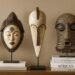 Three Traditional African masks, including a Punu mask, a Fang mask and a Kifebwe mask on stands, displayed on a wooden sideboard with stacked books titled "African Masks." A vase with white roses is on the left.