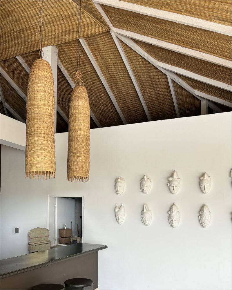 Interior shot of a home showcasing a bar area with two woven pendant lights, eight face sculptures on the wall, and a unique thatched roof. Two large, cylindrical pendant lights woven from a light brown material hang from the thatched roof. The roof itself is intricately designed with diagonal thatched patterns, supported by white beams. Below the lights, set against a white wall, are eight face sculptures arranged in two rows of four. Each sculpture is a simple, white mask-like representation of a human face, each adorned with different headwear or hairstyles. To the left of the sculptures is a bar area featuring a dark gray countertop, two stools, and a glimpse into another room through a doorway. Inside the door, there is a small, light green chair.