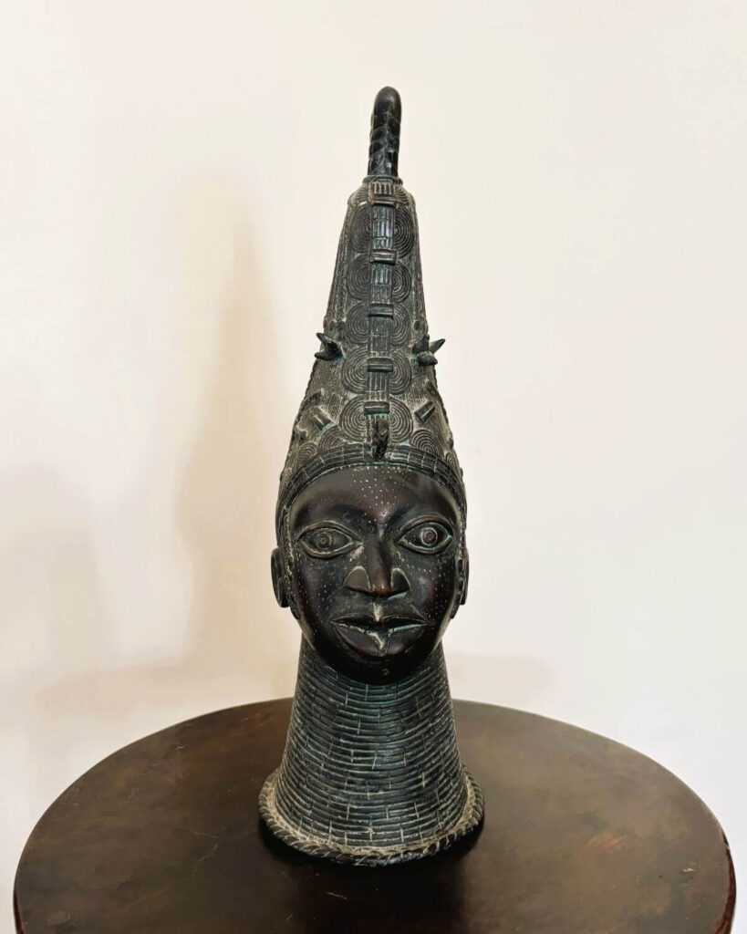 Bronze head of a royal figure standing on a circular wooden table. The bronze head has a flat face with large almond eyes and full lips. The head tapers gradually into a tall conical headdress. The headdress is intricately designed with repeating geometric and linear patterns. Extending upward from the rear of the headdress is a horn-like element that curves backwards, suggesting a stylized representation of a braid or plait. Below the face the neck is textured with closely stacked, horizontal lines resembling stacked rings. The bronze patina has green undertones. The dark wooden table is a complementary piece with its shiny polished surface. A neutral background provides a simple backdrop.