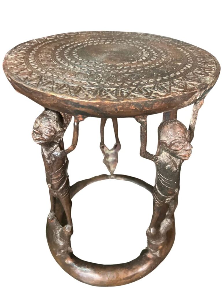 A carved wooden stool. The stool has a round top that is intricately carved, and the legs are made from human figures. The legs connect to a u-shaped bottom that supports the seat.