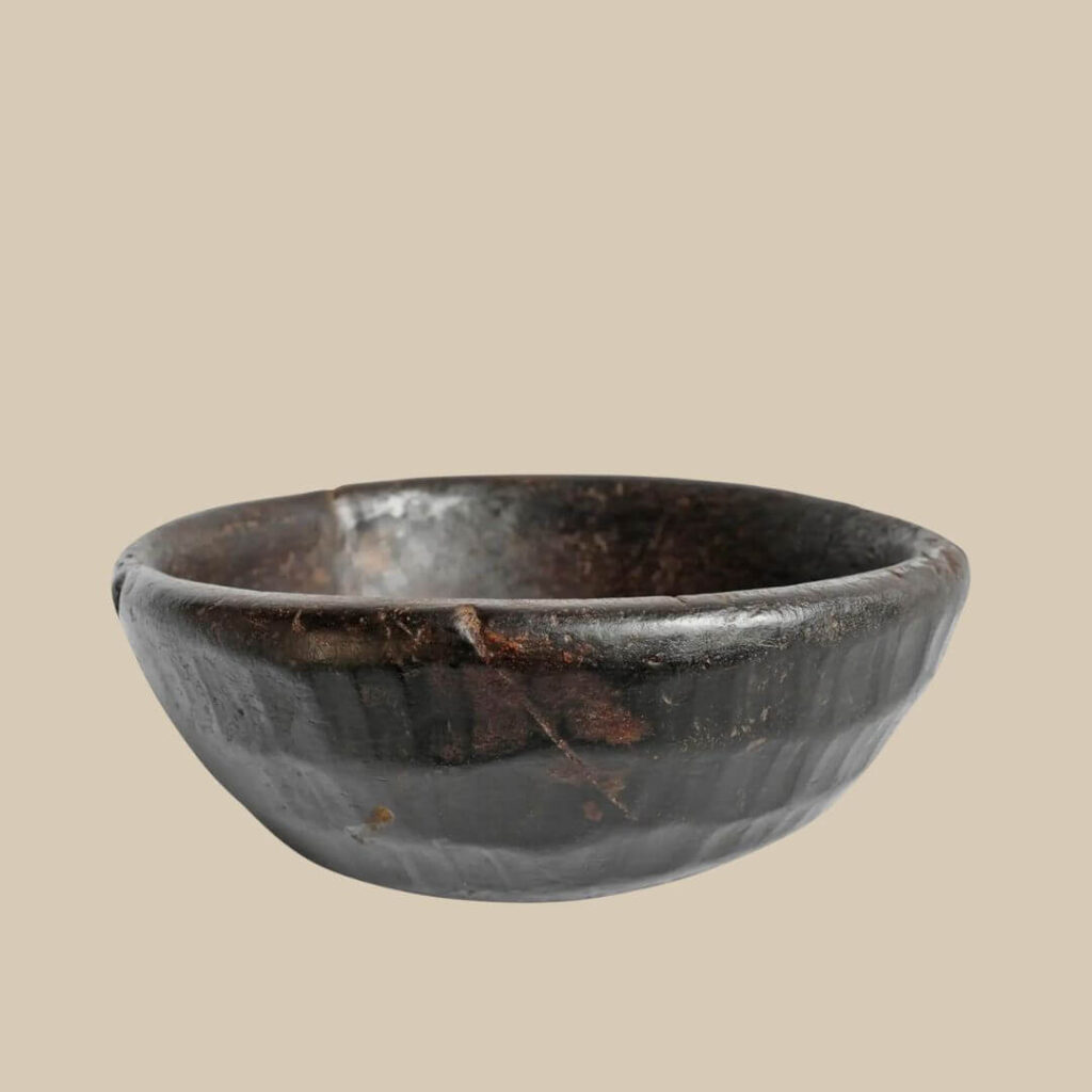A darkened, rustic wooden African bowl sits centered against a light beige background. The bowl's surface is uneven, showing ridges and variations in color from dark brown to reddish hues, suggesting its handcrafted nature and age.