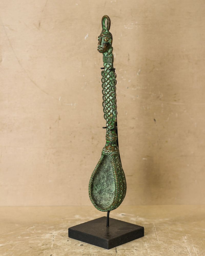 Close-up of a bronze decorative spoon with an intricate long handle and a small square base, set against a plain off-white background. The spoon has a dark patina, with green coloration in the recesses, and is mounted on a black display stand. The handle of the spoon is decorated with a repeating geometric pattern, and the end of the handle features a small human head figure with distinctive facial features.