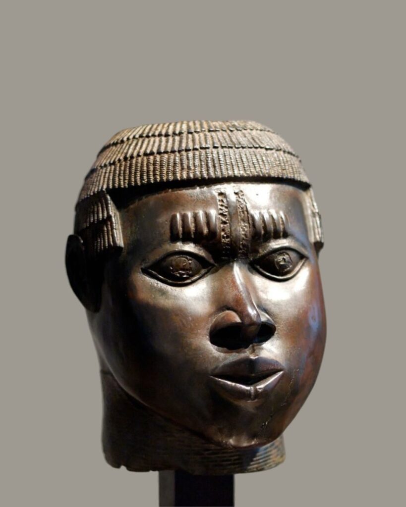 Bronze head of an African man. The statue is set against a blurry gray background and the sculpture is slightly aged and darkly colored. The man has a thick brow, big eyes, a wide nose with large nostrils, and thick lips. His hair is neatly and uniformly braided in thick rows from front to back, with a line of braids on his forehead. There is a single, thin line extending from his hairline down the middle of his forehead.