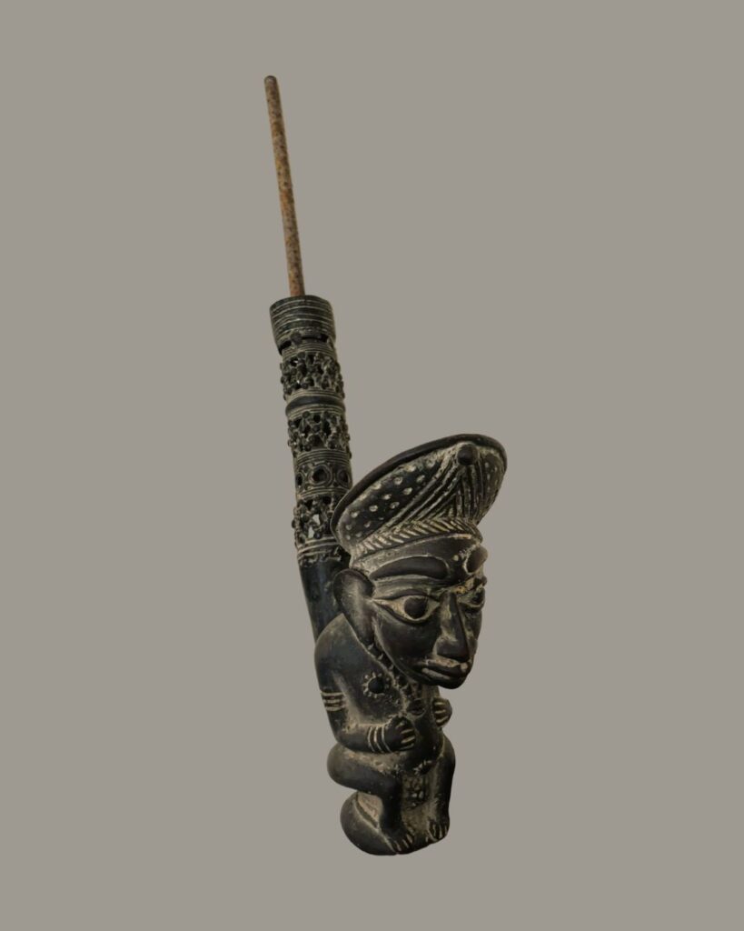 A close-up shot shows an African figurine with intricate carvings set against a plain gray backdrop. The figurine is dark in color and made in bronze. It depicts a person wearing a hat with a high crown. The hat and the surrounding area are detailed with small carvings. The figurine's face features prominent eyes, a defined nose, and a slightly protruding mouth. The person is depicted seated, hugging their knees with their arms. A string with a circular pendant hangs around their neck. Attached to the top of the figurine is a shaft. The base of the shaft leading up from the figurine is decorated with circular carvings with intricate designs within them. A plain metal rod extends upward from the carved section of the shaft.