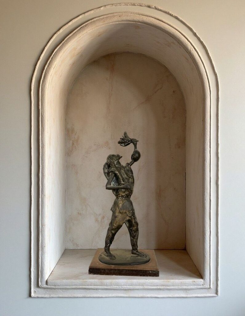 Detailed bronze art sculpture of a figure carrying objects, displayed within an arched wall niche.