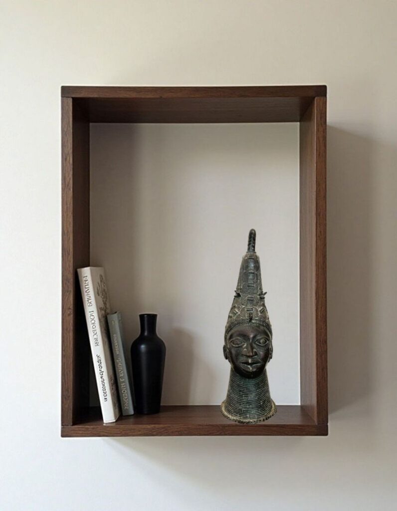 A wooden shelf against a white wall displays three items: two books, a decorative black matte vase, and a striking piece of bronze art—a head sculpture reminiscent of Benin bronzes.
