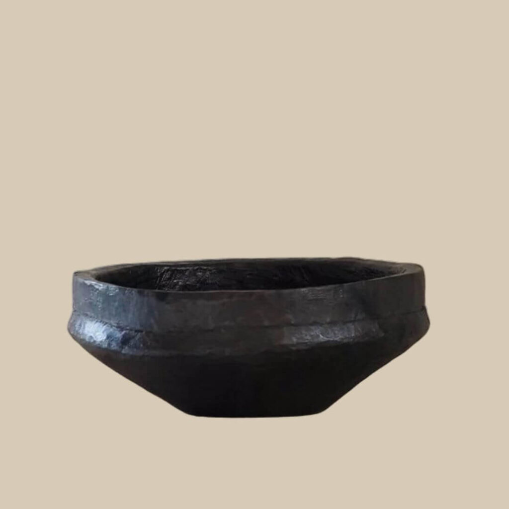 Close-up studio shot of a small, dark, handmade African bowl on a beige background.