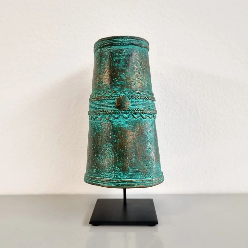 A verdigris-covered bronze bell sits on a black stand, against a white wall. The bell is cylindrical at the top and slightly flared at the bottom. It has decorative bands, including one ornate band with a circular button-like protrusion near the top. Verdigris mottles its surface, giving it shades of green, over top of the brown bronze. The stand holding the bell is a simple, square, black platform with a thin vertical support post. The display surface is light gray. The wall in the background is white.