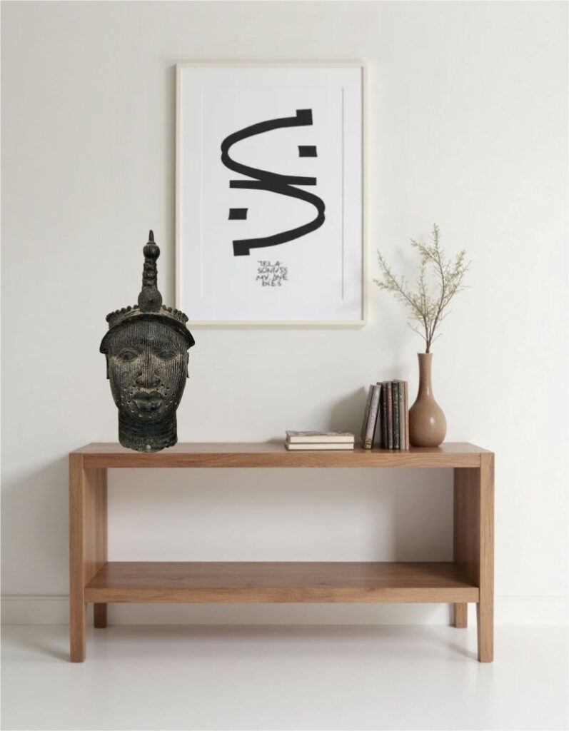 Close-up of a bronze art sculpture of a head on a wooden table against a white wall with a framed art print and a vase of foliage.