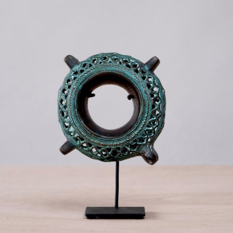 A decorative bronze sculpture is displayed on a black metal stand atop a beige surface set against a neutral gray background. The sculpture features a ring-shaped design with an open center, adorned with intricate latticework around its circumference. Four tubular extensions protrude from the ring at equidistant points. The bronze has a weathered, greenish patina, giving it an antique appearance. The sculpture is centered on the stand, which has a simple, modern design.
