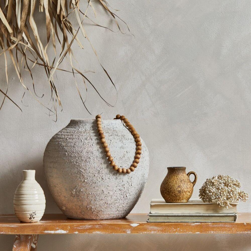 A eye-level medium shot depicts a variety of ceramic vases of differing sizes on top of a wooden ledge. The backdrop features a light gray wall, some dry, straw-like leaves, and dry bunch of small cotton-like plants. On the ledge is a small, plain, white vase that is fluted, a very large, round, textured-gray vase with a rounded beaded necklace coiled around it, a small brown mug behind stacked books and the dry bunch of small cotton-like plants. The ledge appears to be made of a light, naturally-colored, unfinished wood. The light allows the colors of each vases to pop out and the gray-like background color helps it look like a minimalistic scene.