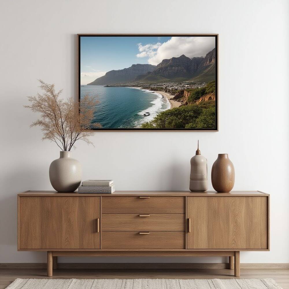 A framed landscape photograph hangs on a white wall, above a wooden sideboard. The photograph depicts a scenic coastal view with mountains rising in the background and a turquoise sea meeting the shore. Puffy white clouds accent the blue sky. Below the photograph, the sideboard is styled with a minimalist aesthetic. On the far left, a round, matte vase holding dried floral stems sits atop two neatly stacked books. To the right are two decorative vases with neutral tones. The sideboard appears to be made of light-colored wood, featuring clean lines, a cabinet door on either side, and three drawers in the middle, with simple, matte gold rectangular handles. The sideboard stands on similar toned solid wood legs. A neutral-colored rug peeks into the frame at the bottom, adding a touch of texture to the otherwise smooth and clean setting. The composition is simple and modern, blending natural elements with contemporary decor.