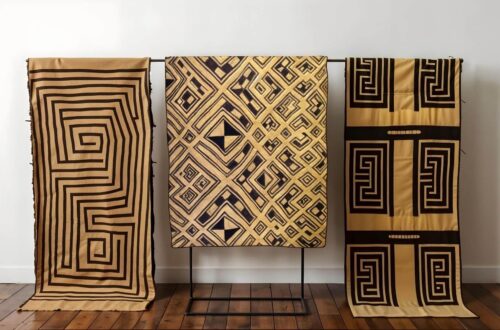 Three panels of geometrically patterned 'Kuba cloth' hang on a metal rack in front of a white wall. The Kuba cloth panels are tan with brown tribal-style designs and hang suspended from a black metal bar. The cloth on the left features a continuous square spiral pattern. The center cloth has a series of nested squares and rectangles, some containing solid black diamond shapes. The cloth on the right utilizes a 'maze' pattern in blocks separating by brown lines. A dark wood floor spreads out below the cloths.