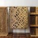 Three panels of geometrically patterned 'Kuba cloth' hang on a metal rack in front of a white wall. The Kuba cloth panels are tan with brown tribal-style designs and hang suspended from a black metal bar. The cloth on the left features a continuous square spiral pattern. The center cloth has a series of nested squares and rectangles, some containing solid black diamond shapes. The cloth on the right utilizes a 'maze' pattern in blocks separating by brown lines. A dark wood floor spreads out below the cloths.