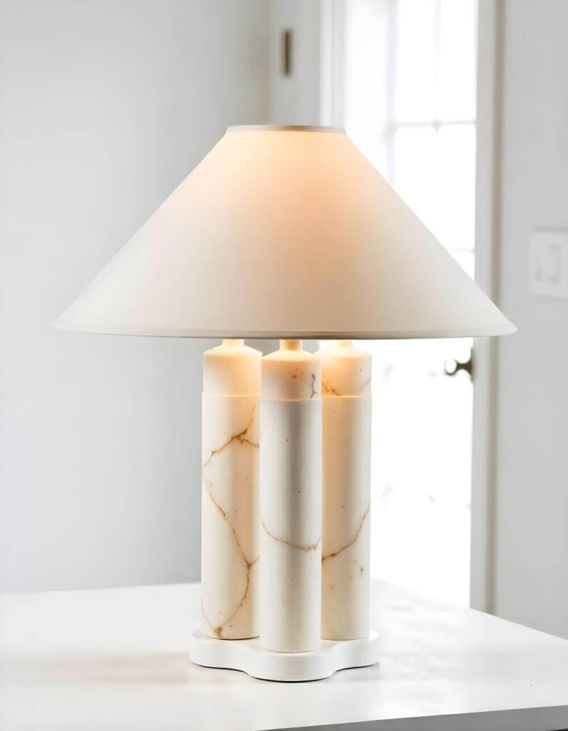 A table lamp with a white shade sits on a white table. The lamp base consists of three cylindrical pillars with a distinctive marble pattern of veined gold. The lamp is lit, casting a soft glow. In the background, a door and wall are softly visible, maintaining the white color scheme.