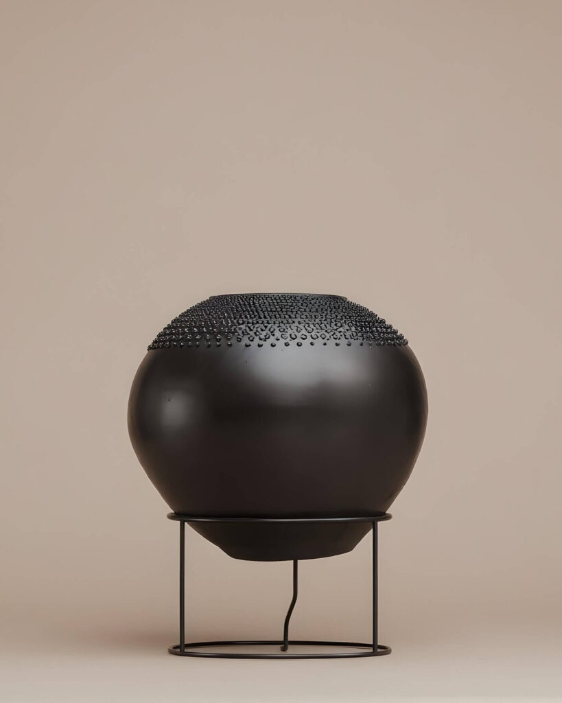 A black spherical vase with textured top sits on a minimal black metal wire stand, all against a light peach-tone background. The vase has rows of small, raised dots or bumps adding a tactile dimension to its upper portion. The stand consists of straight vertical supports connected to circular rings at the top and bottom, providing a simple yet elegant base for the vase. The background helps to highlight the vase and stand.