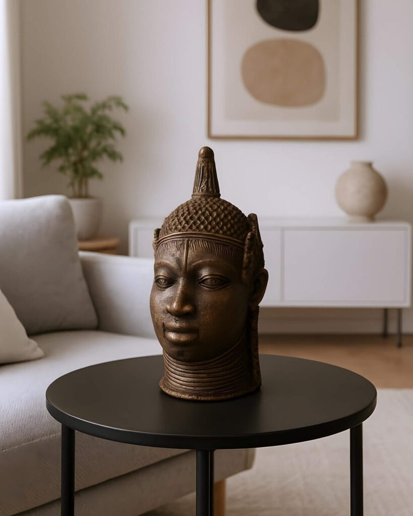 A bronze head sculpture rests on a round black table in a modern living room. The sculpture, reminiscent of Nigerian art, features a detailed representation of a human head, complete with a regal headdress and multiple bands around the neck, suggesting a person of high status. The head's patina suggests age and cultural significance. The table is simple in design with a smooth, matte surface and slender metal legs. It stands on a light-colored rug and is positioned in front of a neutral-toned couch with a pillow. The background reveals a contemporary living space, including a white cabinet, an abstract art piece on the wall, a decorative vase, and a potted green plant. The lighting is soft, highlighting the texture of the sculpture and the sleek lines of the furniture.