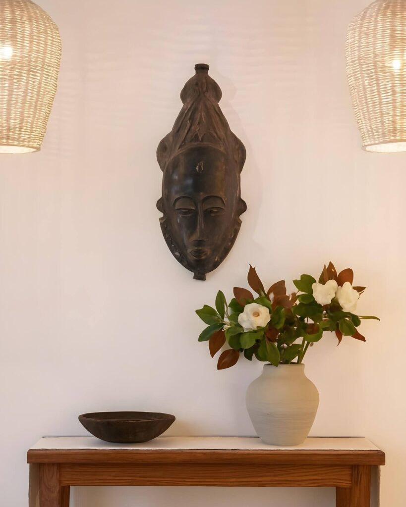 A warmly lit, eye-level shot captures a curated interior design vignette. A wooden table with a white top serves as the base for the arrangement, providing a platform for a simple, dark bowl on the left and a light-colored, full vase on the right. The vase is filled with an assortment of lush green foliage and delicate white flowers, infusing a natural element into the space. Above the table, mounted on the pale wall, a dark, intricately carved African mask adds a cultural or artistic touch. Two pendant lamps with a woven, wicker-like texture flank the mask, casting a warm, inviting glow that ties the composition together.