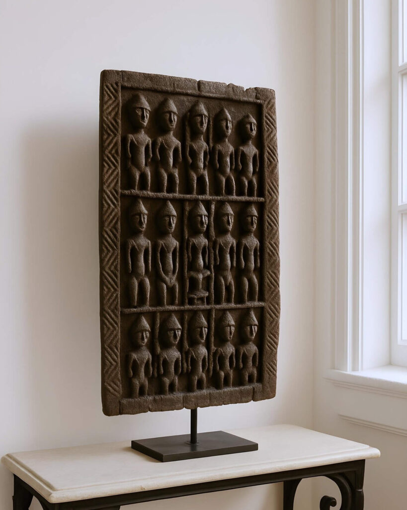 A rectangular carved Dogon door panel depicting eighteen human figures arranged in three rows of six, displayed on a sleek, slender metal stand atop a console table near a window. The stone, likely sandstone or a similar textured material, exhibits a weathered, dark brown hue, adding to its antique or tribal art aesthetic. Each small figure is rendered in a stylized, rudimentary form, featuring tapered heads, arms, and legs, carved in relief. The vertical orientation of the piece is emphasized by the slim metal stand, which is secured to a square base, providing stability and a modern contrast. The console table below has a light-colored marble or stone top with visible veins and a streamlined black base. A geometric zigzag pattern frames the border of the rectangular stone. The piece sits near a large window with multiple panes and white trim, which diffuses soft, natural light onto the scene. The backdrop is a plain white wall.