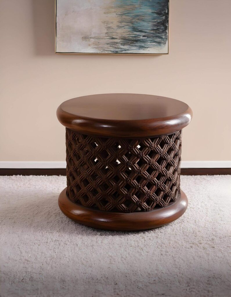 A dark wood end table with a decorative carved drum-shaped base on a cream-colored carpet is a piece of unique home accents. Above the table is a framed piece of artwork.