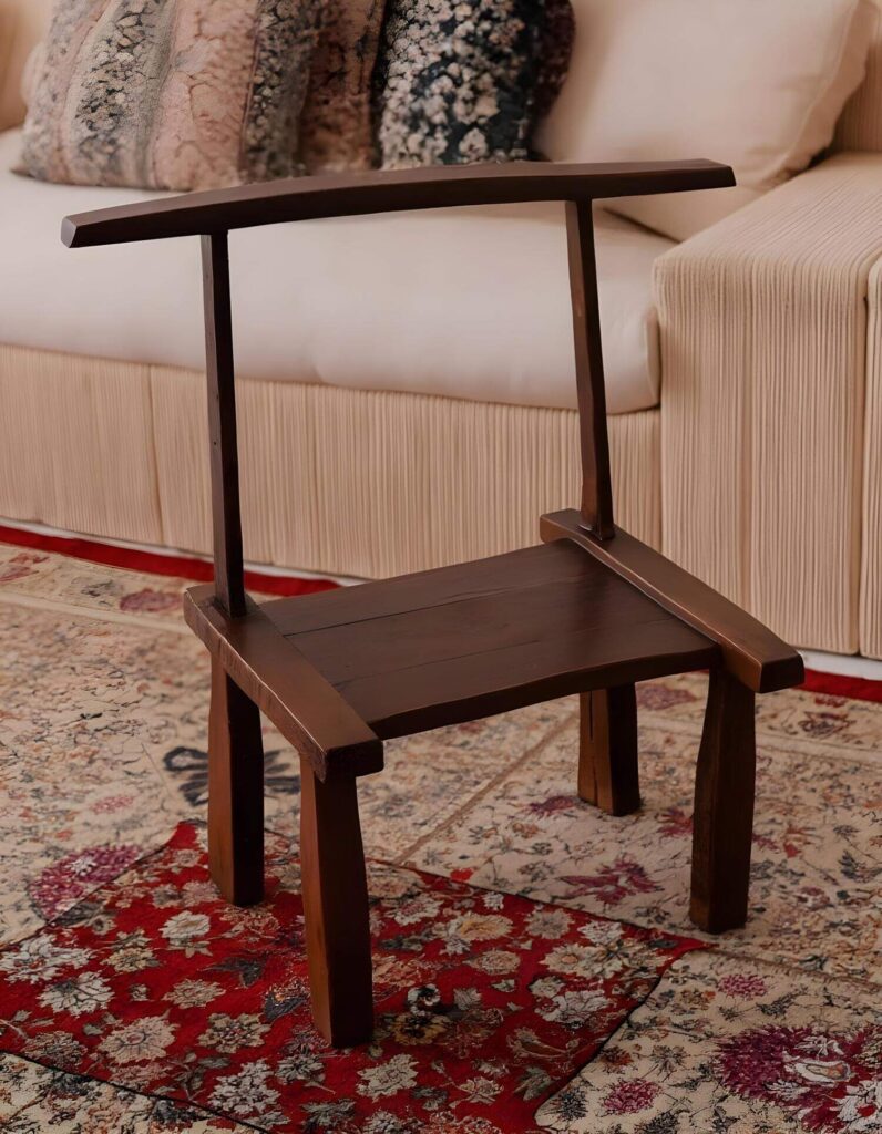 A unique home accent, this dark wood chair with a high back sits on a carpet with red floral design. The simple, angular chair complements a modern interior design aesthetic. In the background is a couch with white cushions.