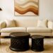 A modern living room scene features a high-end cream sofa, a red velvet armchair and a deep olive-green armchair, flanking a pair of dark, intricately carved Bamileke stools used as coffee tables, all set on a cream-designer rug, with a framed abstract art piece hanged above the sofa.