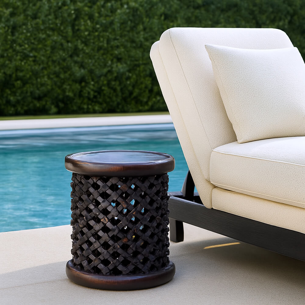 A Bamileke stool sits on a patio next to a white cushioned lounge chair; behind the chair is a bright blue pool.