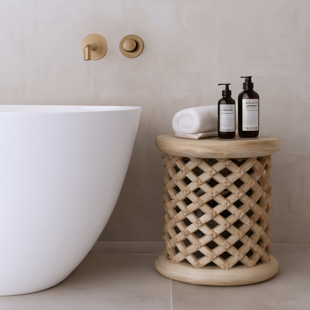 A decorative Bamileke stool sits beside a modern white tub. The light wood stool is topped with a white, rolled up towel and two pump bottles. Gold faucet fixtures are mounted on the wall behind the tub. Neutral walls and flooring create a soothing, spa-like atmosphere.