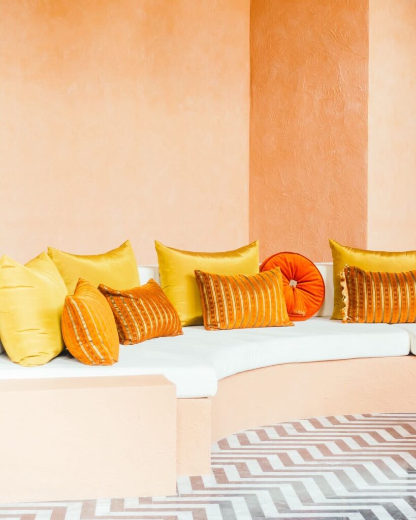 Moroccan interior design featuring a built-in white sofa against an orange textured wall, adorned with yellow and orange throw pillows, set on a geometric patterned tile floor.