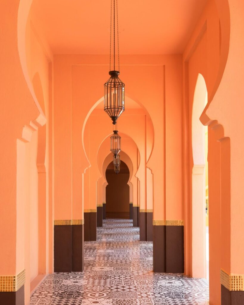 An eye-level shot captures a symmetrically designed hallway with a series of archways, progressing into the distance. The walls are coated in a warm, peachy orange hue, contrasted by the dark brown bases of the pillars that support the arches. The floor is adorned with a patterned tile design in black and white and a band of gold, mosaic-like tiles decorates the lower sections of the walls and pillars. Elegant, wrought iron lanterns dangle from the ceiling at intervals.