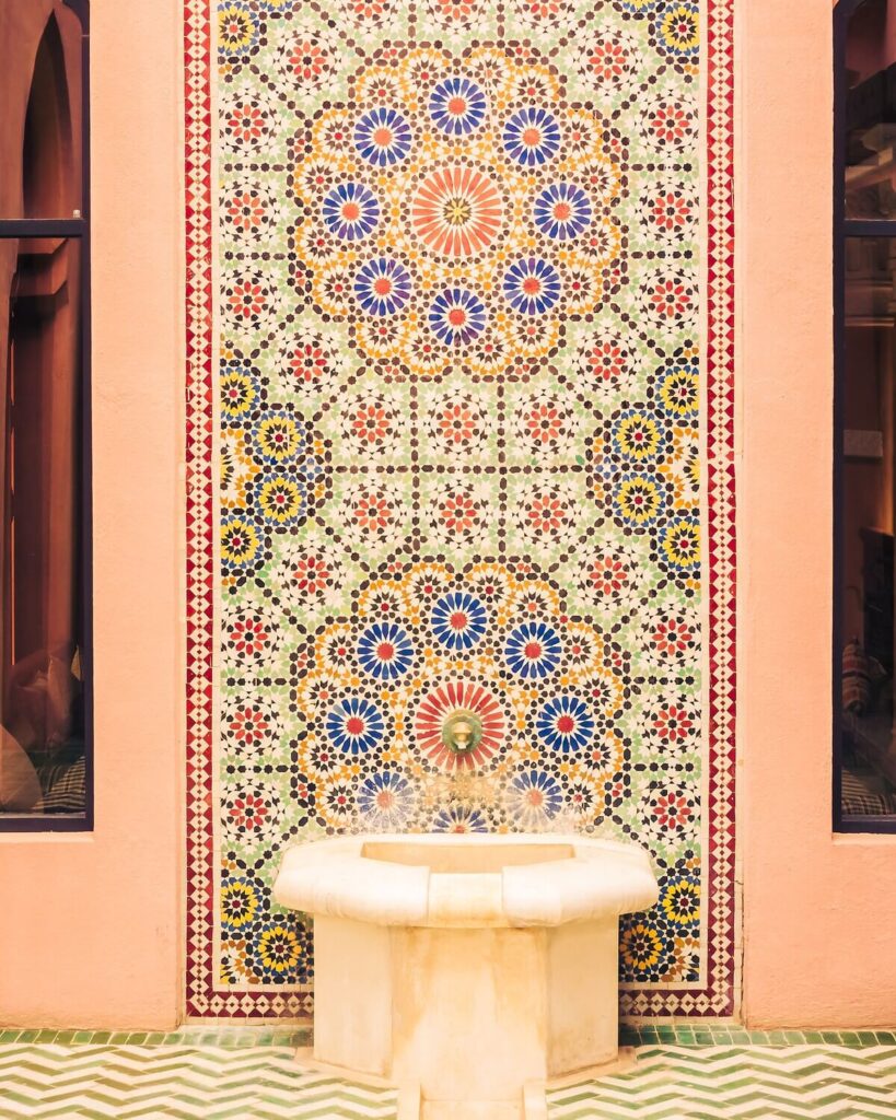 A wall fountain is set against a backdrop of intricate Moorish tilework. The fountain is a simple, white stone structure with a shallow basin. Water trickles from a small, metallic nozzle embedded in the tilework above. The tilework features a complex geometric pattern with colorful floral motifs in shades of blue, red, yellow, and green. The surrounding walls are painted in a warm, light pink, and dark-framed windows flank the tiled panel. The floor is covered in a green and white herringbone pattern, completing the courtyard's vibrant and culturally rich ambiance.
