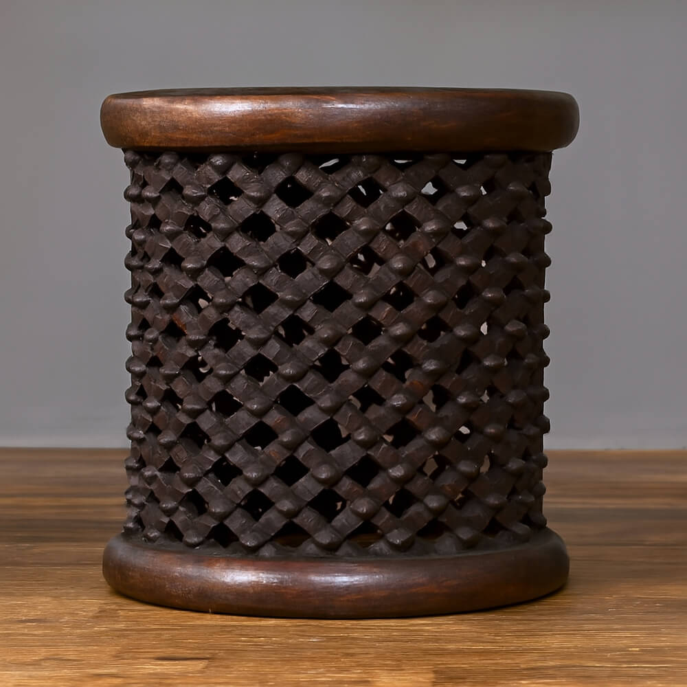 A dark wooden Bamileke stool sits on a warm wooden floor against a plain gray background. The cylindrical stool features a detailed lattice-like pattern with small studs adorning each intersection. It is topped and bottomed with smooth, rounded bands of richer toned wood.