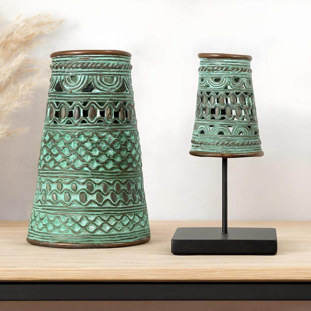 Two decorative African bronze cuffs with a patina and intricate patterns sit on a wooden surface. One is on a black stand, creating a stylish, earthy, and artistic look.