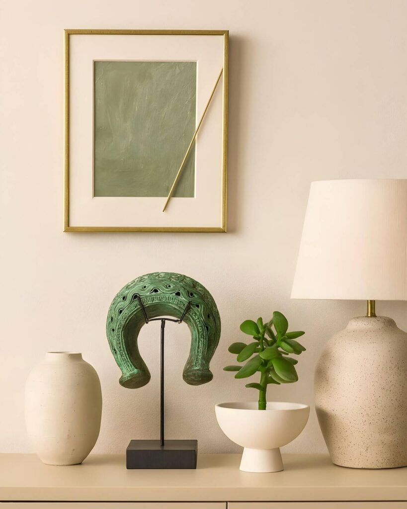 A minimalist arrangement on a light-toned surface features a thick Benin bronze bangle with a patina, a jade plant in a white bowl, ceramic vases, a table lamp, and abstract wall art.