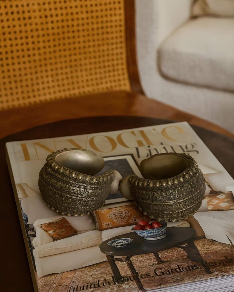 Two ornate bronze bangles rest on a stylish living coffee table book atop a wooden table. A chair with woven cane back and a white sofa set the cozy tone.