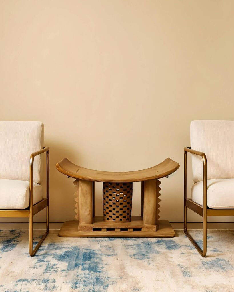 Light wood Ashanti stool, the most regal of all African stools in between two modern chairs with cream cushions and golden frames on a designer rug with a cream base and blue tie-dye patterns.