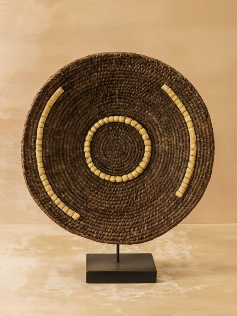 A brown woven Makenge basket with a round, shallow shape, featuring concentric circles of beige, held upright on a black metal stand.