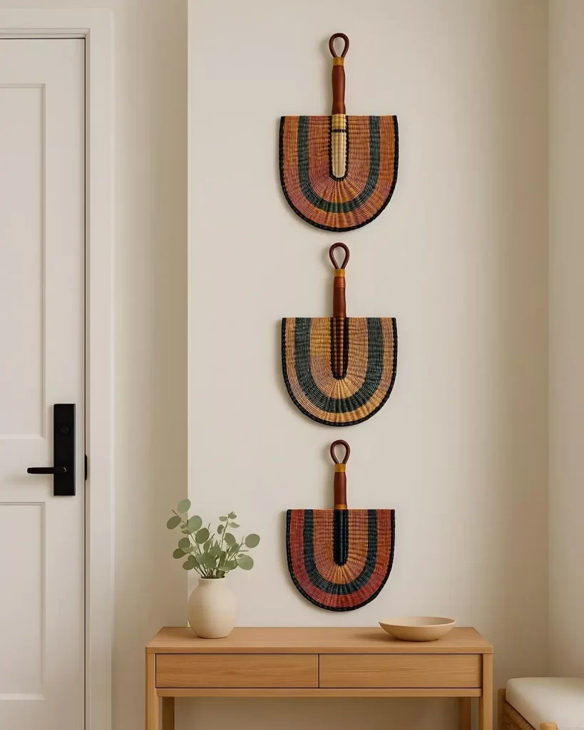 Three matching, semi-circular Bolga fans with woven stripes of red, orange, green, and dark blue, hung vertically on a cream wall above a wooden console table.