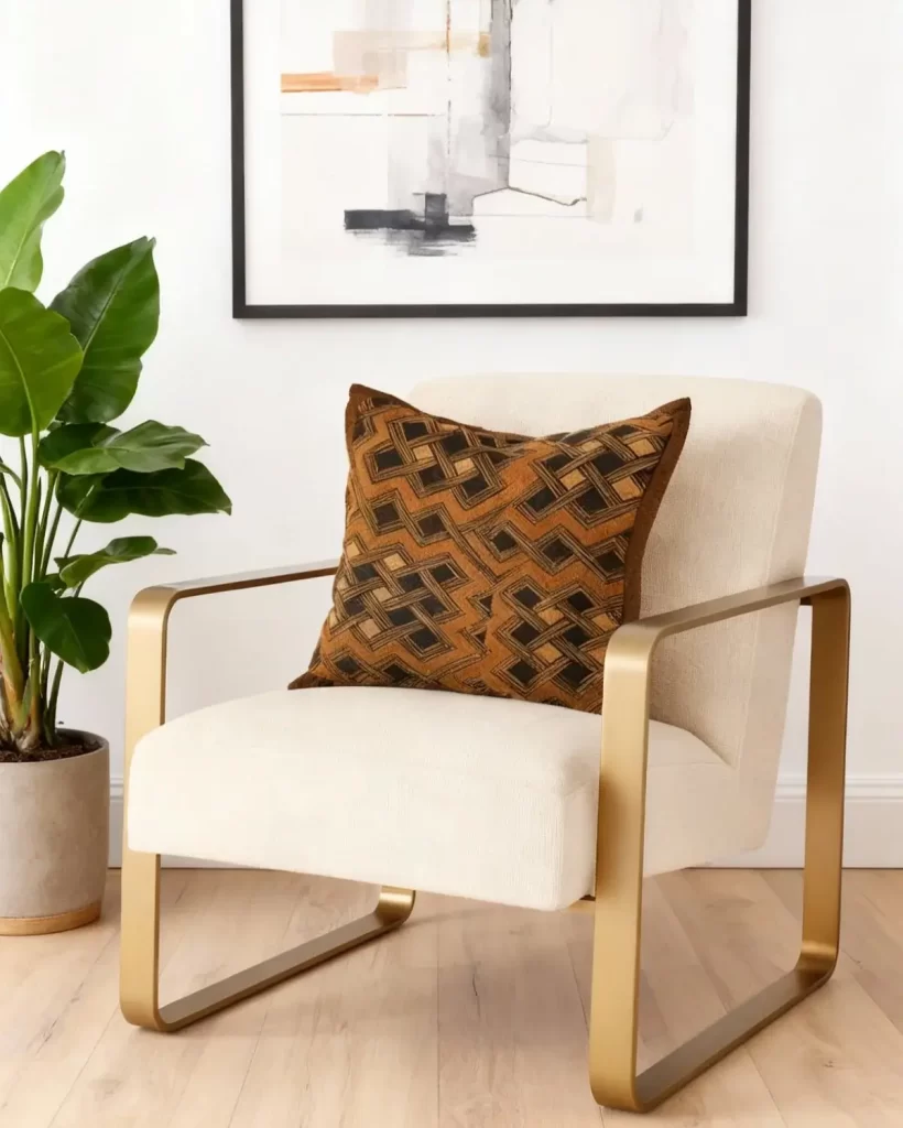 Modern armchair with gold arms and a geometric-patterned Kuba cloth cushion, placed on a light wooden floor. Behind, a potted plant and abstract wall art enhance the cozy atmosphere.