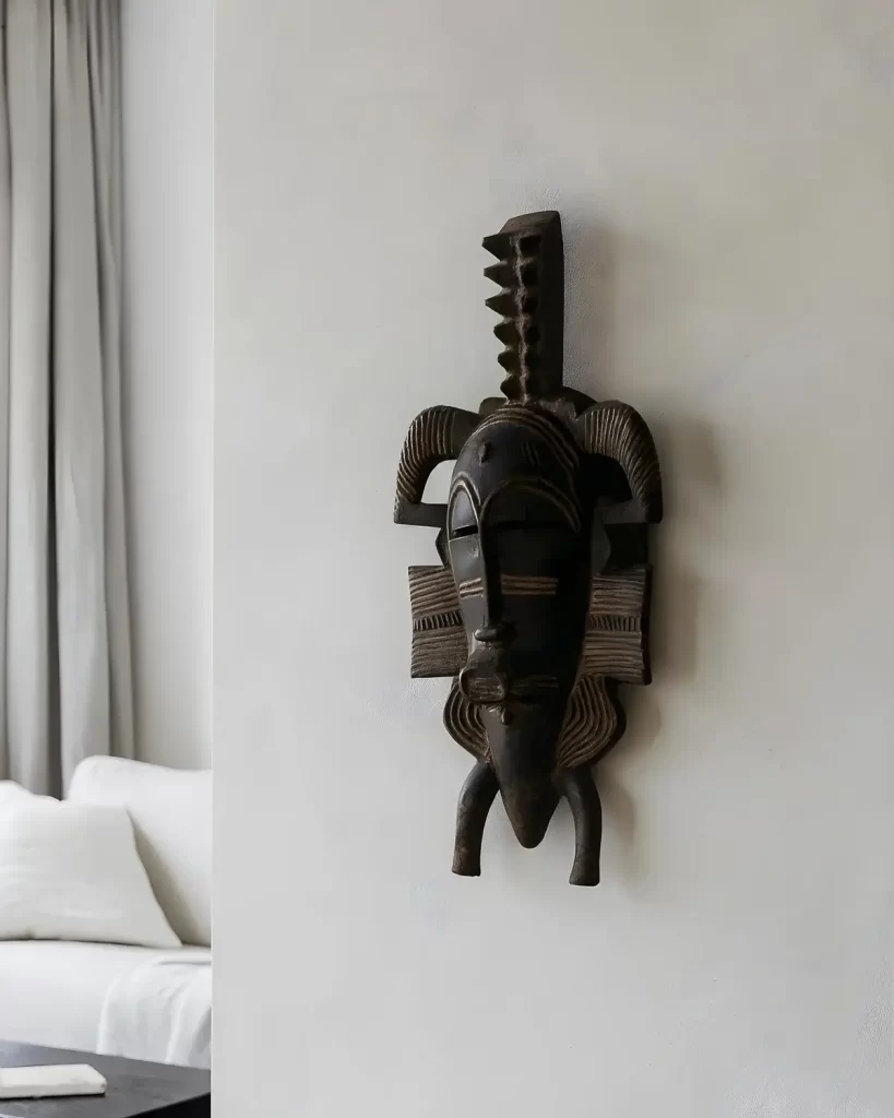 Wooden Senufo mask with intricate carvings hangs on a white wall, conveying a cultural and artistic vibe. Minimalist decor with a white sofa nearby.