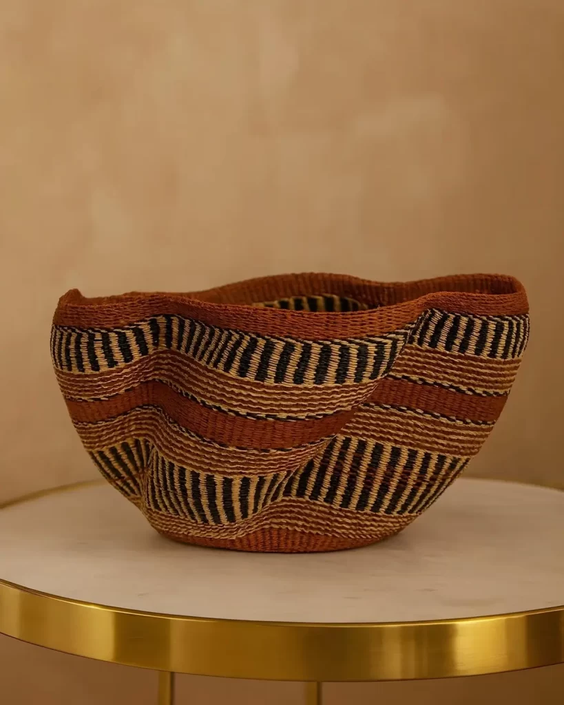 A hand-woven Pakurigo wave basket with an undulating rim, featuring horizontal stripes of black, natural tan, and rusty orange, sitting on a small, round table.